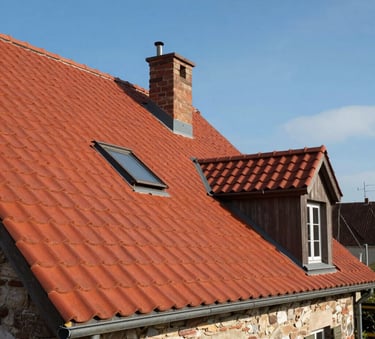 Une photographie architecturale d'une maison de style européen avec un toit en tuiles rouges impeccablement rénové, sous un ciel bleu clair, mettant en valeur la propreté du travail de couverture.