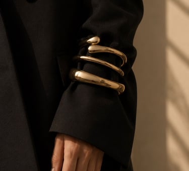 Gold statement bracelets worn over an oversized black blazer sleeve in soft natural light.