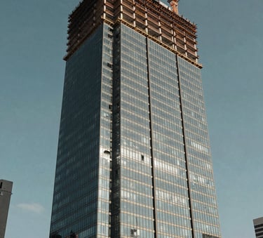 A low-angle high-resolution shot of a modern vertical glass building under construction with a crane visible, in a bustling Latin American / Spanish city, bright daylight, Muted Slate Blue sky, professional photography.