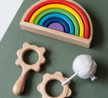 A flat-lay photograph of curated children's developmental toys, including a wooden rainbow stacker and a soft cotton rattle, arranged neatly on a deep sage green surface with white accent elements. Clean, modern aesthetic.