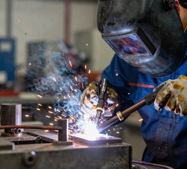 A close-up photograph of a professional industrial welding process in a Brazilian workshop, intense golden and blue light from the arc, a worker in protective gear partially visible in a soft focus background, slate blue-gray and steel gray metallic tones.