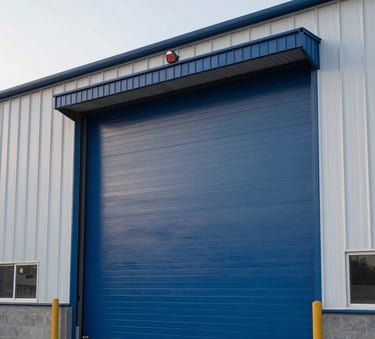 A wide shot of a modern industrial warehouse entrance featuring a large steel blue power door, soft pearl white sky in the background, professional architectural photography.