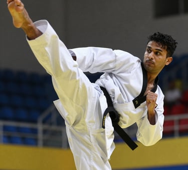 A close-up, high-action photograph of a South Asian martial artist's legs mid-air performing a powerful side kick. The athlete wears a clean white Taekwondo dobok with a prominent black belt. The background is a modern, slightly blurred sports hall with dark blue and yellow accents, focusing on the precision and intensity of the movement.
