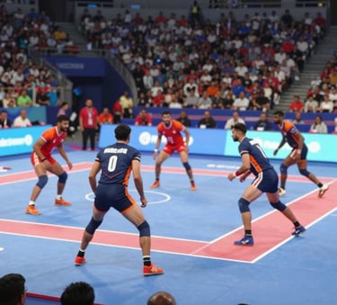 A dynamic wide-angle shot of a Kabaddi match in a brightly lit stadium in Brampton, Ontario. The image captures the energetic crowd and players in action, featuring the brand's deep blue and orange-red colors in the team uniforms. Professional sports photography style.