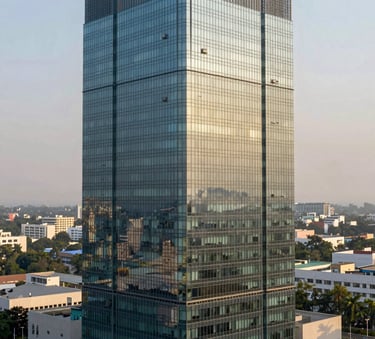 A high-angle architectural photograph of a modern glass-clad corporate tower in a South Asian / Indian business district like Gurugram. The building reflects a clear sky, and the surrounding environment includes landscaped greenery in shades of Muted Sage Green and Light Grey-Green. Professional lighting during the golden hour.