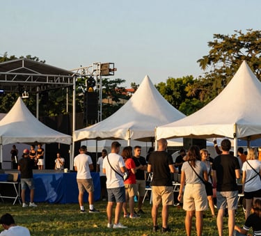 A vibrant photo of a community event in a local park. People are gathered near a steel blue stage. The lighting is warm golden hour sunlight, reflecting off soft off-white event tents.
