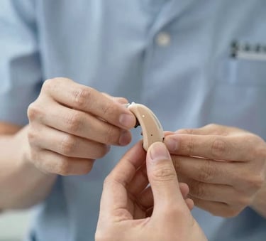 A close-up shot of a professional audiologist's hands showing a tiny, discreet hearing aid to a patient. The focus is on the device's design and accessibility. Lighting is bright and trustworthy, using the #1A2C38 brand color in the uniform and #A7C9D6 in the background accents.