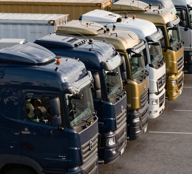 A fleet of commercial transport trucks and water bowsers lined up perfectly in a logistics hub in the Gulf, photographed at a professional side angle, featuring deep slate blue and metallic gold details on the vehicles.