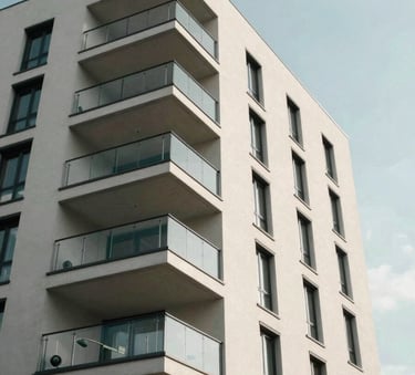 A low-angle shot of a modern apartment building in Berlin under a clear sky. The architecture features clean lines, glass balconies, and a facade in soft off-white and muted sage tones, conveying stability and professional management.