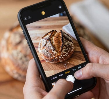A close-up shot of a digital marketing specialist's hands holding a smartphone, capturing a high-quality photo of a rustic sourdough bread on a wooden surface (#4A3C32). Warm, natural lighting with soft shadows and hints of Crimson (#9B1C1C) in a nearby napkin.