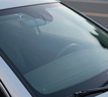 A detailed close-up photograph of a pristine, newly installed windshield on a luxury car. The glass is perfectly transparent, reflecting a soft blue North American sky. The setting is clean and modern, focusing on the high-quality finish and clarity.