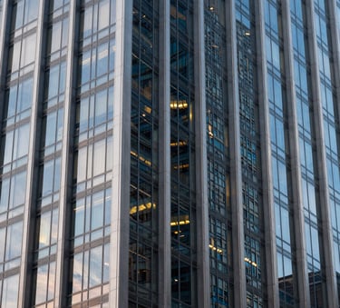 A close-up of a contemporary glass and steel skyscraper facade in a US city at dusk, showing light grey and dark blue reflections with sharp architectural focus.