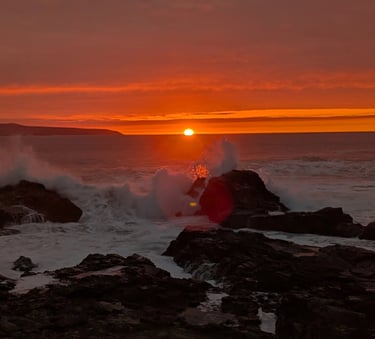 Golden sunset over the ocean with waves crashing against rocky coastline under an orange sky.