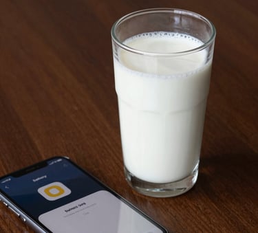 A close-up photograph of a fresh glass of milk on a deep espresso brown wooden table next to a smartphone showing a delivery notification. The lighting is clean and minimalist.