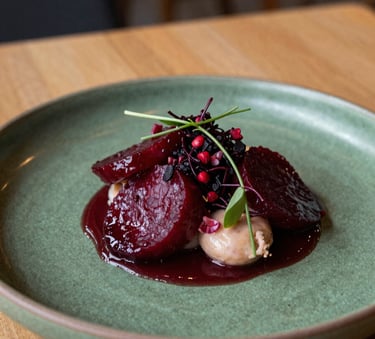 A close-up photograph of a beautifully plated artisanal dish in a cozy Northern European / Scandinavian restaurant, featuring vibrant deep ripe crimson ingredients on a matte forest green ceramic plate under warm, natural lighting.