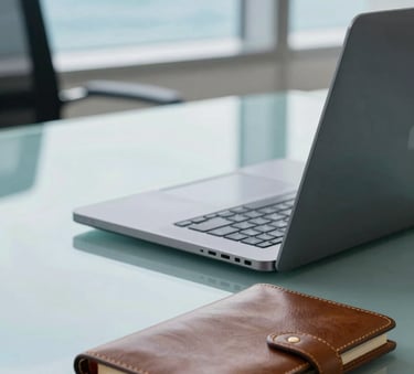 A close-up of a high-end laptop and a leather notebook on a glass desk in a modern Latin American / Spanish corporate office, with soft focus on ocean blue and pale mist blue interior details, natural bright light.
