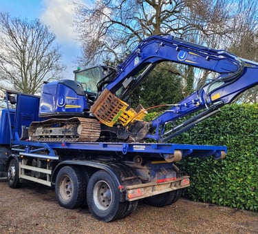 A blue JMC tracked excavator loaded onto a heavy-duty flatbed transport truck parked outdoors.