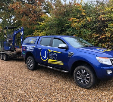 A blue JMC Groundworks Ford Ranger truck towing a mini excavator on a gravel driveway.