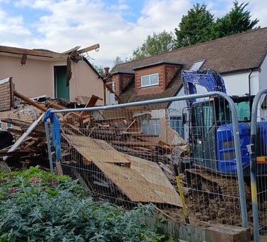A blue excavator demolishing a residential house behind a construction safety fence.