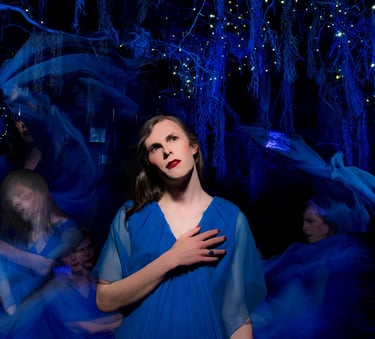 a trans woman in a blue dress with her hand on her heart. There are blurred blue images behind her