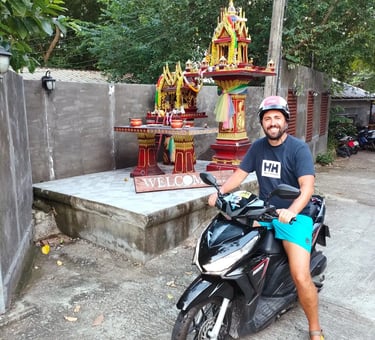 A smiling man wearing a helmet sits on a black scooter in front of a colorful Thai spirit house.