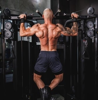 Muscular man performing wide grip pull-ups on a cable machine at the gym to build back muscles.