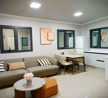 Modern tiny house interior with a grey sofa, dining set, and compact kitchen featuring white cabinets.