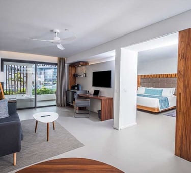 Modern hotel suite with a living area, work desk, and bedroom view through a sliding wooden door.