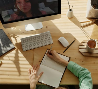 A woman takes notes in a notebook during a remote video conference call on her desktop computer.