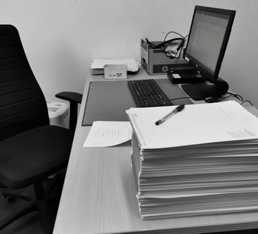 Black and white photo of a workspace with a computer, keyboard, and a large stack of paper documents.