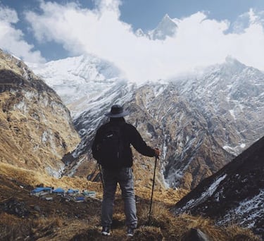 A hiker with trekking poles overlooks a snowy mountain valley in the Himalayas.