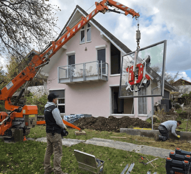 Oranger Minikran hebt mit einem Saugheber eine große Glasscheibe zur Montage in das Obergeschoss eines Wohnhauses.