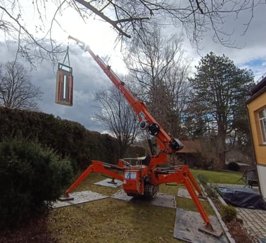 Oranger Minikran hebt ein Bauelement im Garten, stabilisiert durch Stützen auf Bodenschutzplatten.