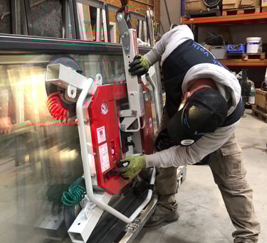 Schreinermeister Ferdinand Volk justiert den roten Vakuum-Saugheber an einer großen Glasscheibe, die an einem Kranhaken ist.