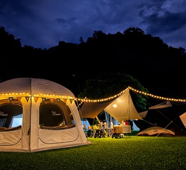 Illuminated camping tents and string lights on a grassy field at night with mountain silhouettes.