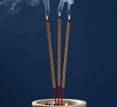 A close-up of incense sticks burning in a ceramic holder, with soft smoke curling upwards against an indigo background, South Asian / Tamil Nadu, India.