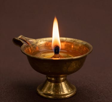 A close-up photograph of a sacred brass oil lamp with a single flickering flame, casting a warm golden glow against a dark brown background. South Asian spiritual aesthetic.