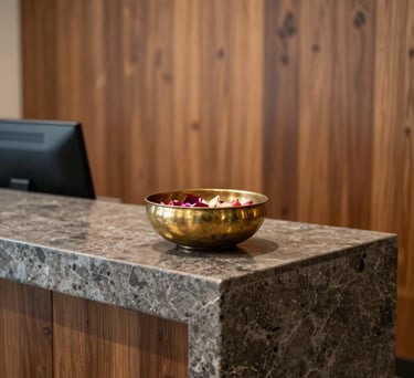 A serene office reception area with a modern stone desk, a South Asian decorative brass bowl with floating flower petals, and warm wooden wall panels.