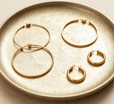 A beautifully arranged flat lay photograph of multiple minimalist gold jewelry pieces, including delicate bracelets and hoop earrings, on a textured champagne gold ceramic tray. Bright and airy lighting, sophisticated luxury aesthetic, warm beige background.