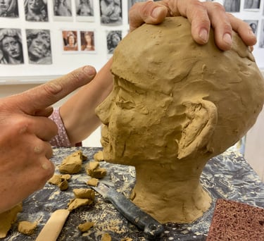 An artist sculpting a clay head bust in a studio with portrait sketches on the wall.
