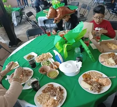 Familia disfrutando barbacoa con arroz y frijoles en una mesa verde durante una fiesta infantil.