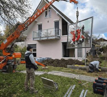 Oranger Minikran hebt mit einem Saugheber eine große Glasscheibe zur Montage in das Obergeschoss eines Wohnhauses.