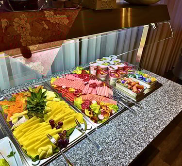 Hotel breakfast buffet featuring platters of sliced cheese, cold cuts, salmon, and yogurt containers.
