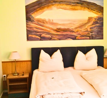 Cozy hotel room with double bed, bedside lamps, and a scenic canyon landscape painting above the headboard.