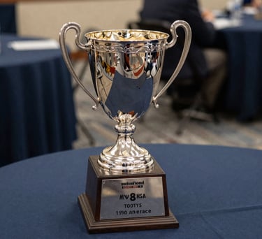 A close-up of a silver championship trophy sitting on a table with a dark navy blue tablecloth. Soft light reflects off the metal. North American / US Southern collegiate athletic banquet setting.