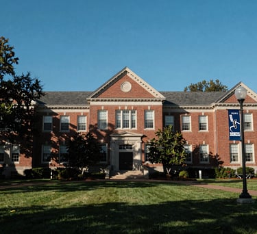 Photography of a large university campus building with traditional Southern architecture, surrounded by green lawns under a clear blue sky. A banner for the hockey program is visible on a nearby lamp post.