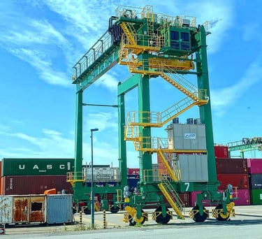 A large green gantry crane standing over shipping containers at a busy commercial port terminal.