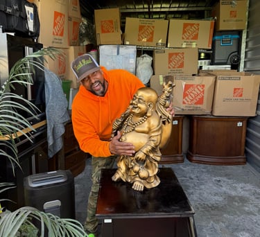 A man in an orange hoodie holds a large golden Buddha statue in a storage unit filled with moving boxes.