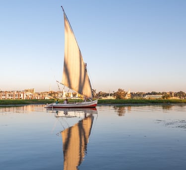 A traditional Egyptian felucca sailboat with a white sail reflecting on the calm Nile River at sunset.