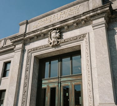 A close-up photograph of the architectural details of the Robert F. Kennedy Department of Justice Building in Washington D.C., focusing on the heavy bronze doors and classic limestone carvings, clear blue sky background, North American / US.